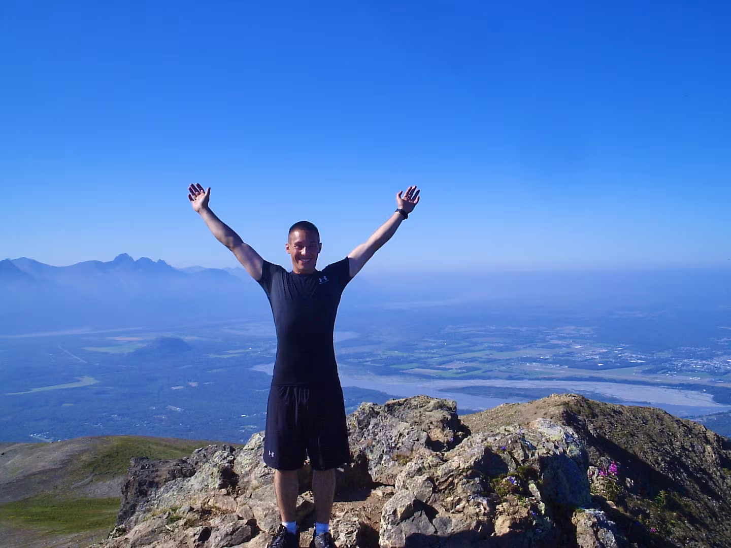 Shane Woods standing on an Alaska mountain summit with his arms raised